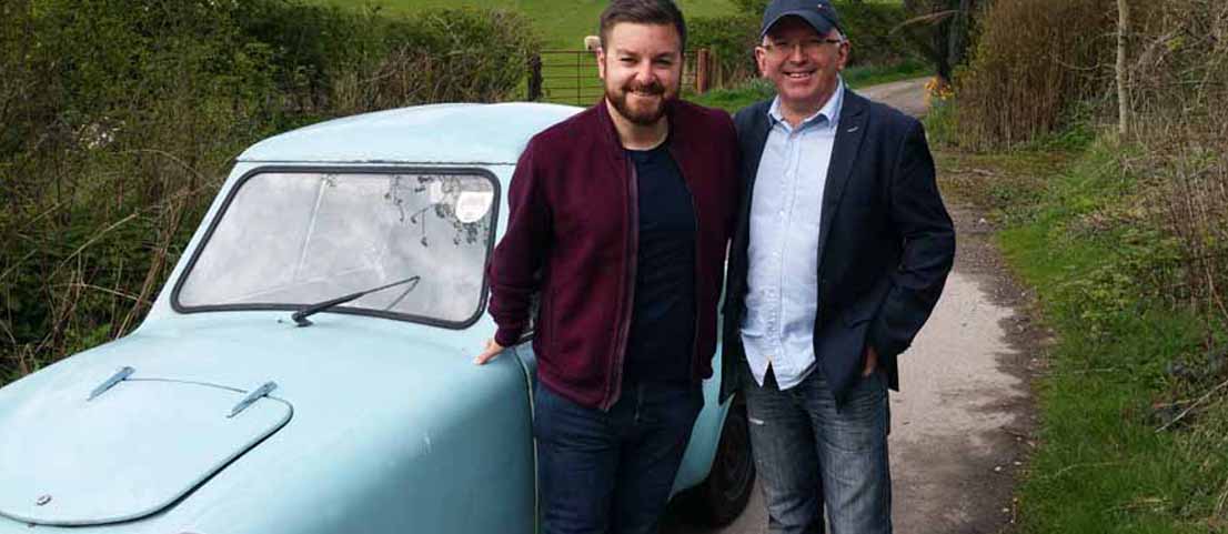 Two men standing beside a pale blue Invacar invalid carriage on a country lane