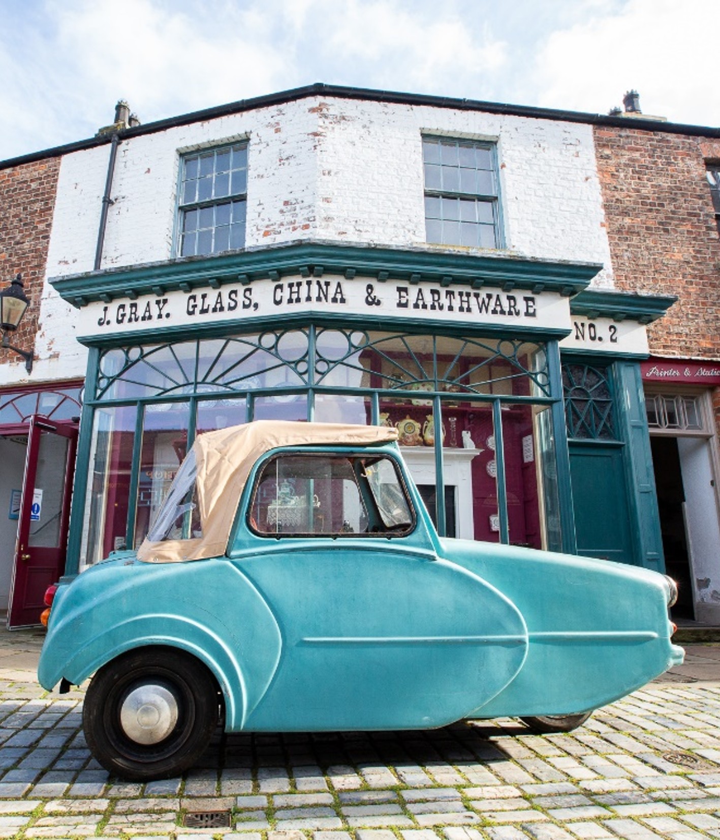Turquoise Tippen invalid carriage displayed outside a historic shopfront at Preston Park Museum.